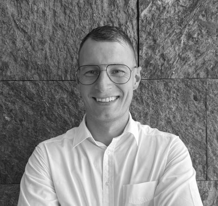 Fabian, il nostro assistente di direzione, è in piedi davanti a una parete nera e sorride alla telecamera con le braccia incrociate.