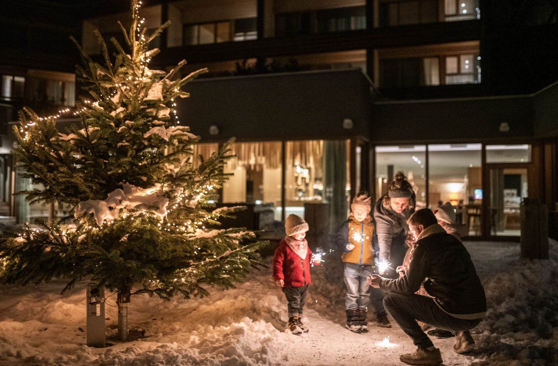 Familie steht vor dem Haus und vor dem Christbaum und zünden einen Sternspritzer an.