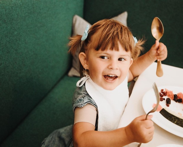 Kleinkind mit Besteck in der Hand und Teller auf dem Tisch