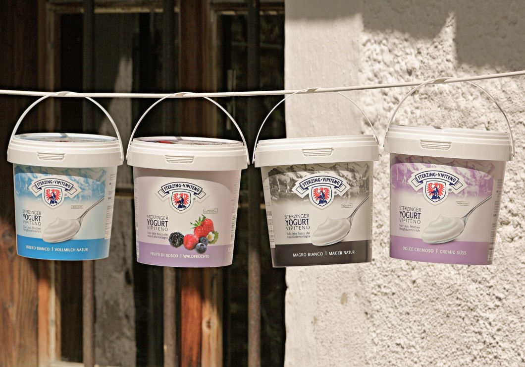 Drei Eimer mit frischer Milch und Joghurt hängen an einer Wäscheleine vor einer rustikalen Wand.