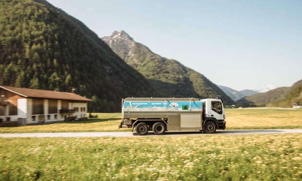 Ein Milchtransporter fährt über eine grüne Wiese mit Bergen im Hintergrund.