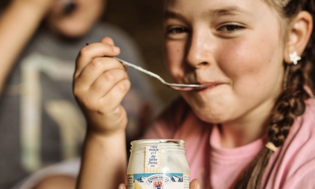 Ein lachendes Mädchen genießt einen Becher Bio-Joghurt und schaut fröhlich in die Kamera.