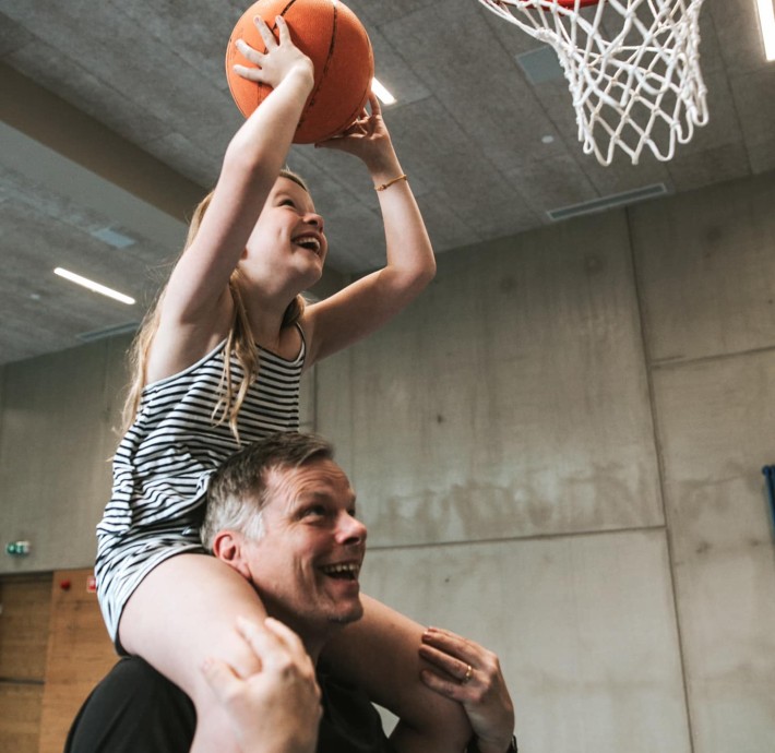 Vater haltet Tochter auf den Schultern, dien ein Basketball in den Korb wirft