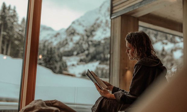 Eine Frau sitzt in einer gemütlichen Fensterbank mit Blick auf die verschneiten Berge.
