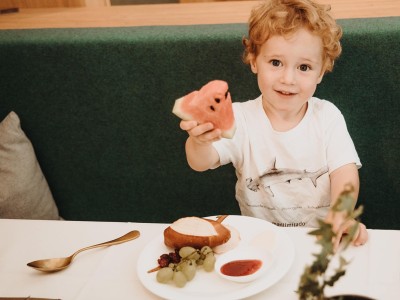Kleines Kind sitz am Tisch und hält ein Stück Wassermelone in die Luft
