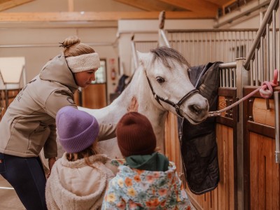 Reitlehrerin und zwei Kinder stehen neben ein junges Pferd, angehängt beim Stall