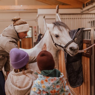 Reitlehrerin und zwei Kinder stehen neben ein junges Pferd, angehängt beim Stall